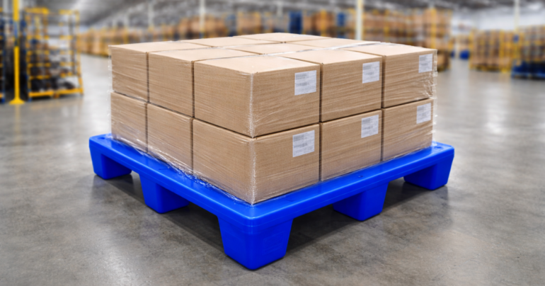 Heavy-duty blue plastic pallet placed on a warehouse floor, loaded with neatly stacked and shrink-wrapped brown cardboard cartons arranged in uniform rows; each carton labeled with white identification tags. The pallet features a raised, reinforced design with multiple block supports for stability and forklift handling. Background shows a clean, organized industrial storage environment with blurred shelving and inventory racks, indicating a logistics or distribution setting.